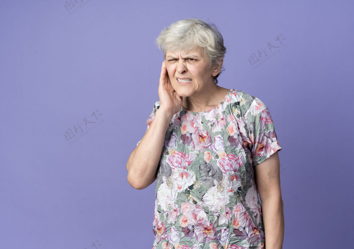 手恼羞成怒的老妇人把手放在隔离在紫色墙上的耳朵上老老烦