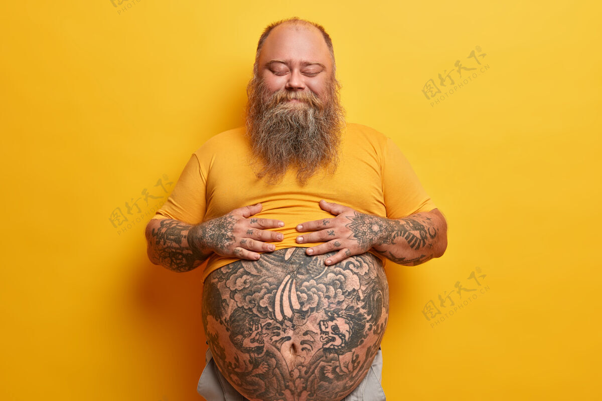 成人喜怒无常的胖男人双手放在肚子上 吃了美味的晚餐后感到饱足 闭着眼睛站着 不在乎身材 荷尔蒙失调 孤立在黄色的墙上大问题脂肪团