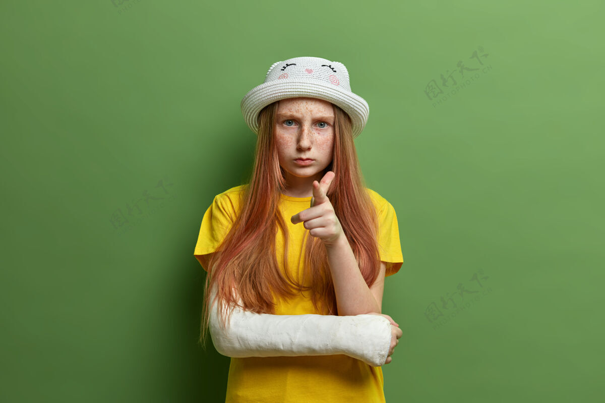 年轻皮肤上有雀斑 长着姜黄色长发的小女孩 指着你严肃地看着你 戴着帽子 穿着黄色t恤 意外摔断了胳膊 被隔离在绿色的墙上显示争论破碎