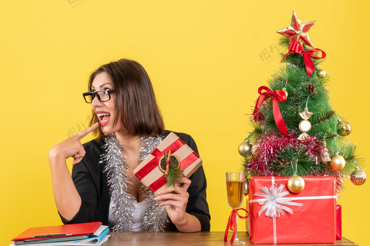 坐着一位穿着西装 戴着眼镜的商务女士拿着礼物 坐在办公室的一张桌子旁 桌上放着一棵圣诞树快乐眼镜漂亮