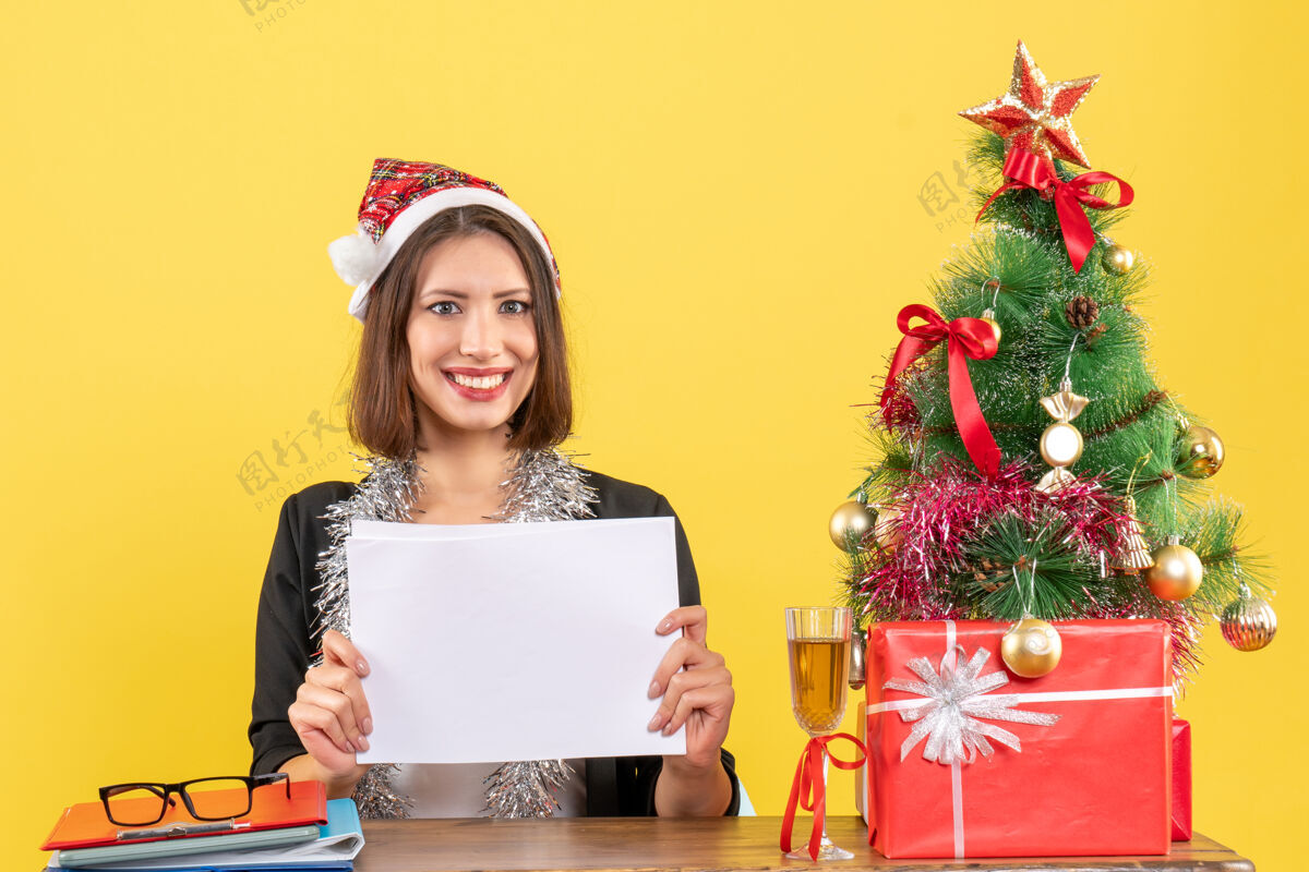 年一位穿着西装 戴着圣诞老人帽 戴着新年装饰品的商务女士独自一人拿着文件 坐在一张桌子旁 桌子上放着圣诞树帽子独自快乐