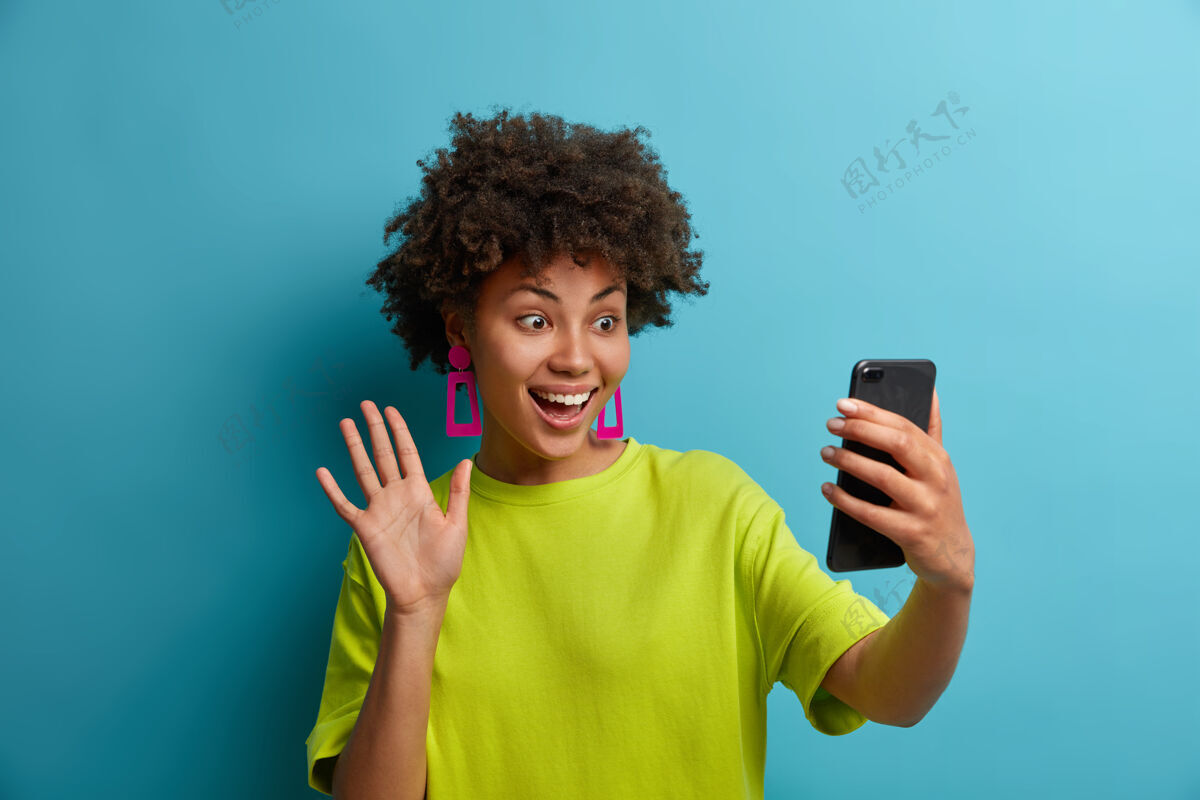 你好快乐的卷发千禧一代女孩在智能手机上自拍 视频通话中有对话 挥舞问候手势 做视频博客广播 表情欢快 与世隔绝非洲流在线