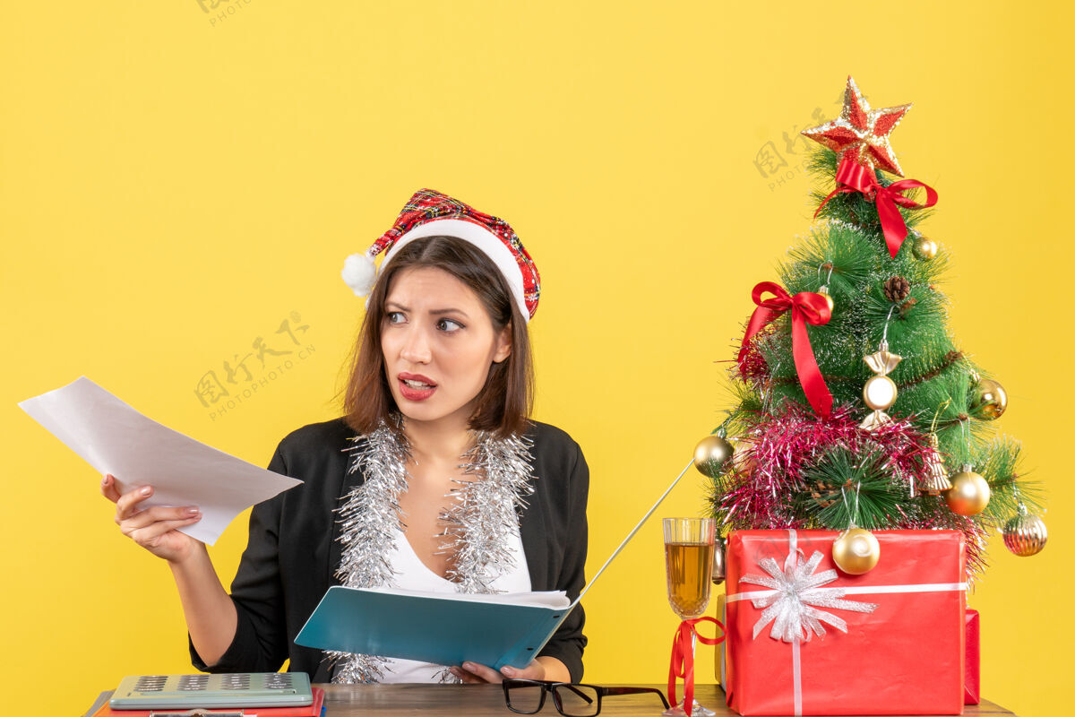 文件好奇迷人的女士穿着西装 戴着圣诞老人的帽子和新年的装饰品 手里拿着文件 在黄色的办公室里与世隔绝圣诞老人人帽子