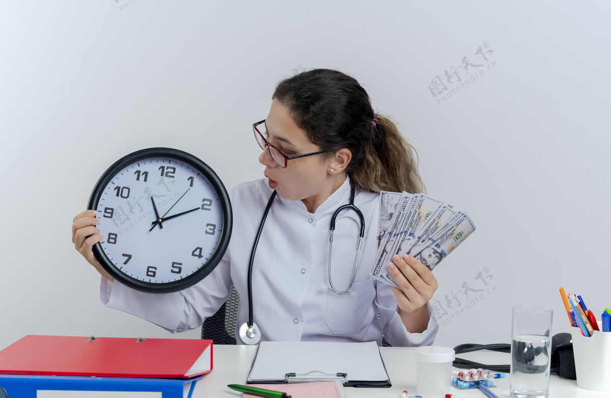眼镜年轻的女医生穿着医用长袍 戴着听诊器 戴着眼镜 坐在办公桌旁 手里拿着医疗工具 手里拿着钱和钟 孤立地看着钟抱着金钱人