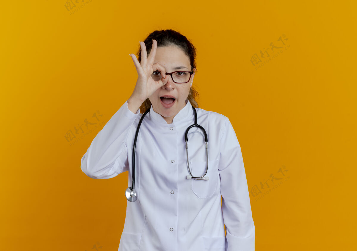 女性快乐的年轻女医生穿着医用长袍 戴着眼镜听诊器 表情孤僻手势欢乐长袍