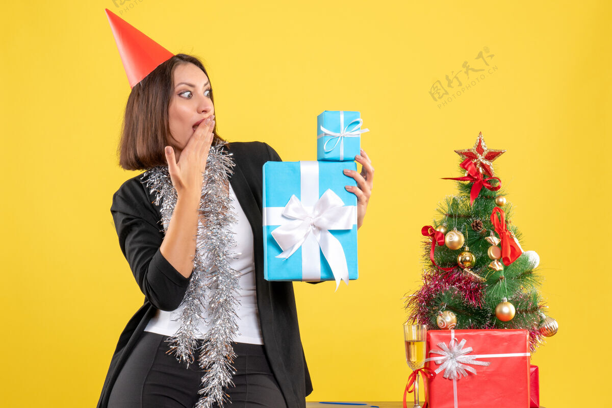 包圣诞节心情与震惊美丽的女士戴着圣诞帽拿着礼物在黄色的办公室人帽子微笑