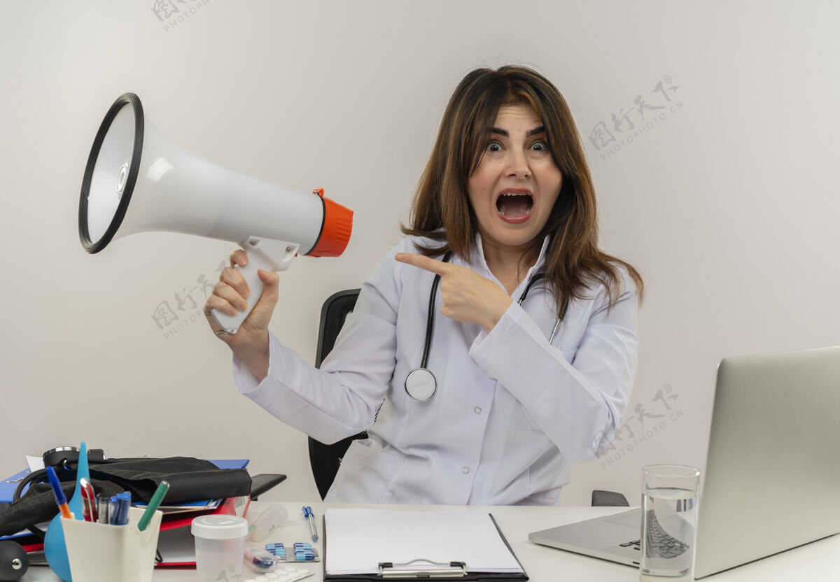 个人令人印象深刻的中年女医生穿着医用长袍和听诊器坐在办公桌旁 带着医疗工具剪贴板和笔记本电脑 拿着扬声器指着隔离的一侧听诊器医疗工具