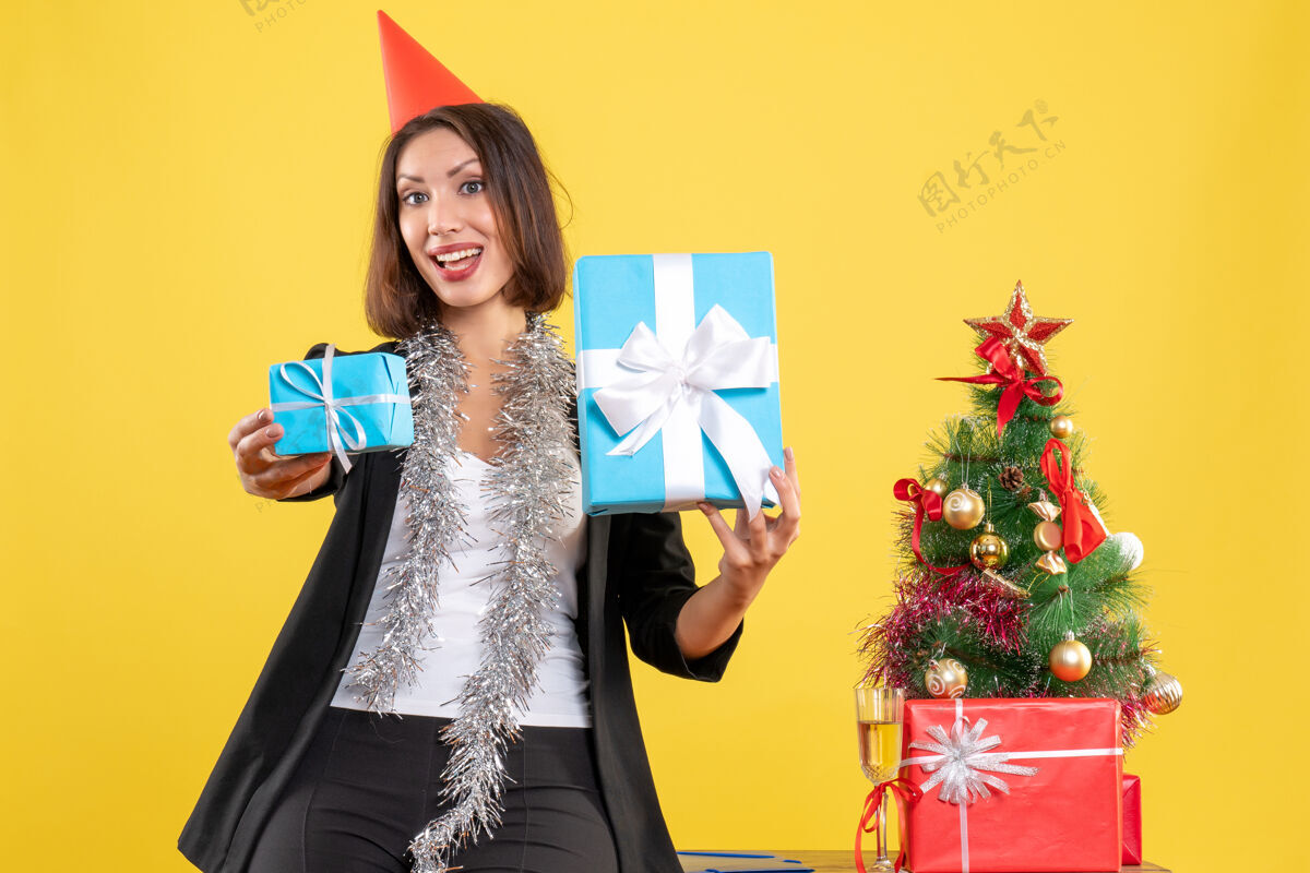 快乐圣诞心情惊喜美丽女士戴着圣诞帽在黄色的办公室里开心地展示礼物黑发模特晚餐礼服
