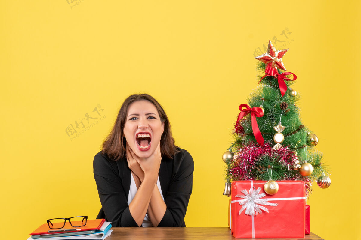 愤怒愤怒和紧张的年轻女子坐在一张桌子旁装饰圣诞树在黄色办公室靠近人坐着