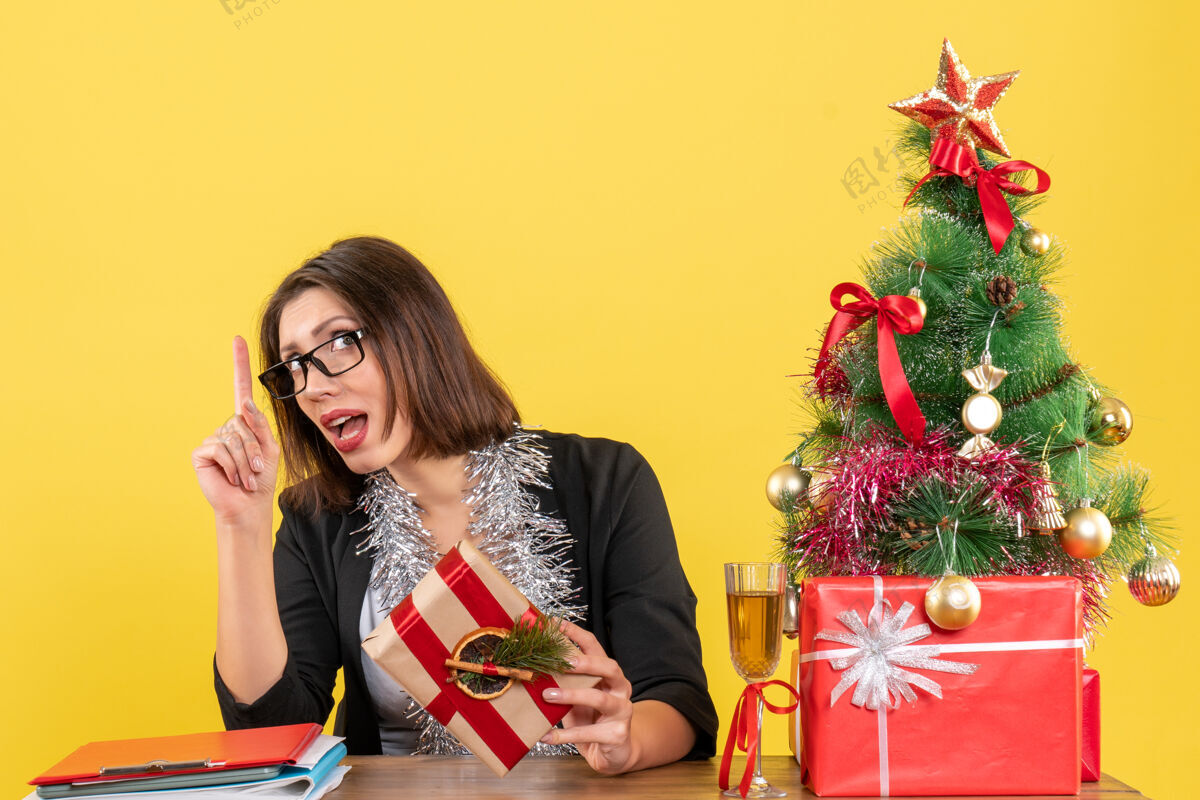指着一位穿着西装 戴着眼镜的漂亮商务女士坐在一张桌子旁 桌子上有一棵黄色的圣诞树眼镜黄色坐着