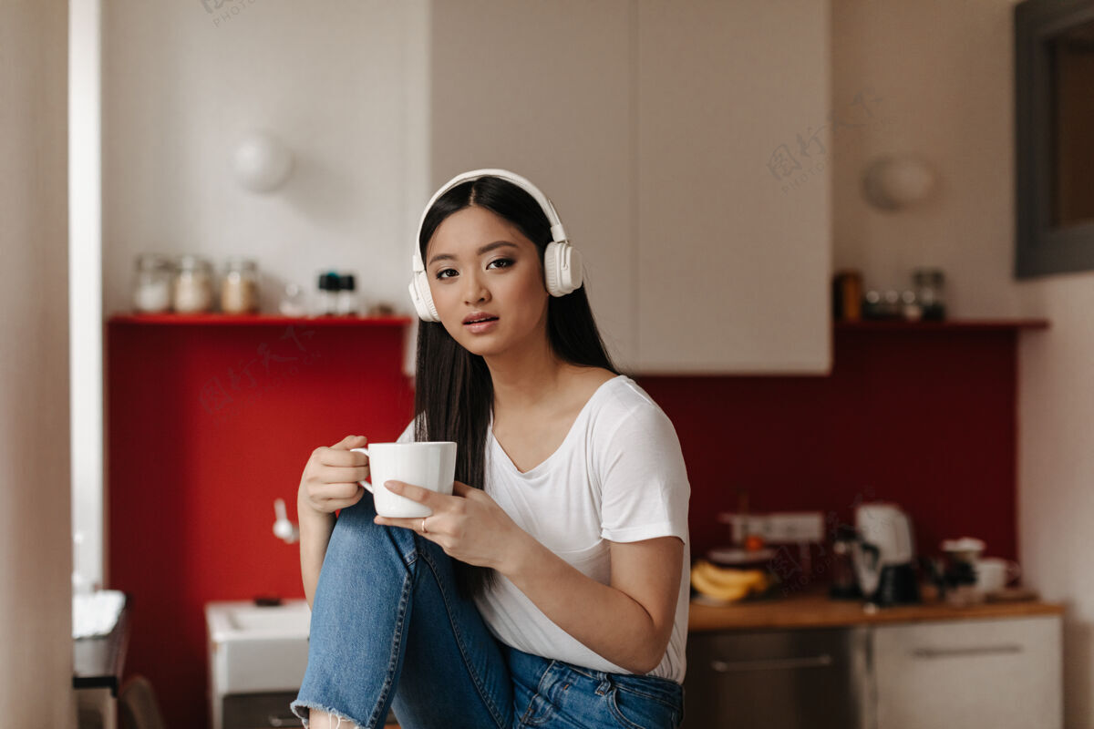 牛仔裤一个棕色眼睛 穿着白色t恤 戴着大耳机的女人看着前面 在厨房的背景上摆着杯子的姿势应用程序沙发搜索