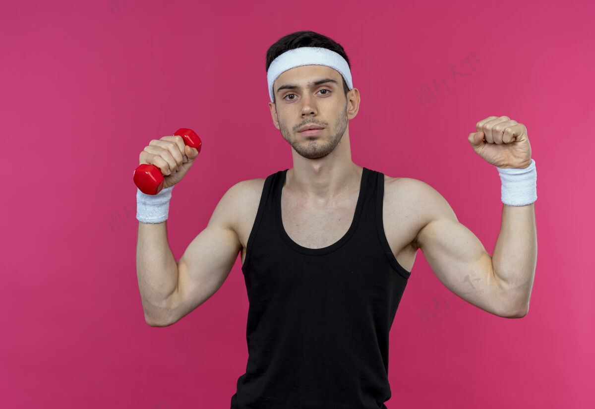 站立戴着头巾的年轻运动型男子 站在粉色背景下 紧张而自信地打着哑铃锻炼身体工作运动自信