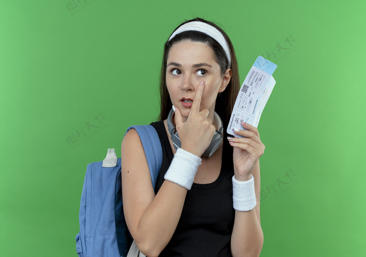 立场戴着头巾 背着背包 拿着机票 手指着眼睛站在绿色背景上的年轻健身女士指向机票背包