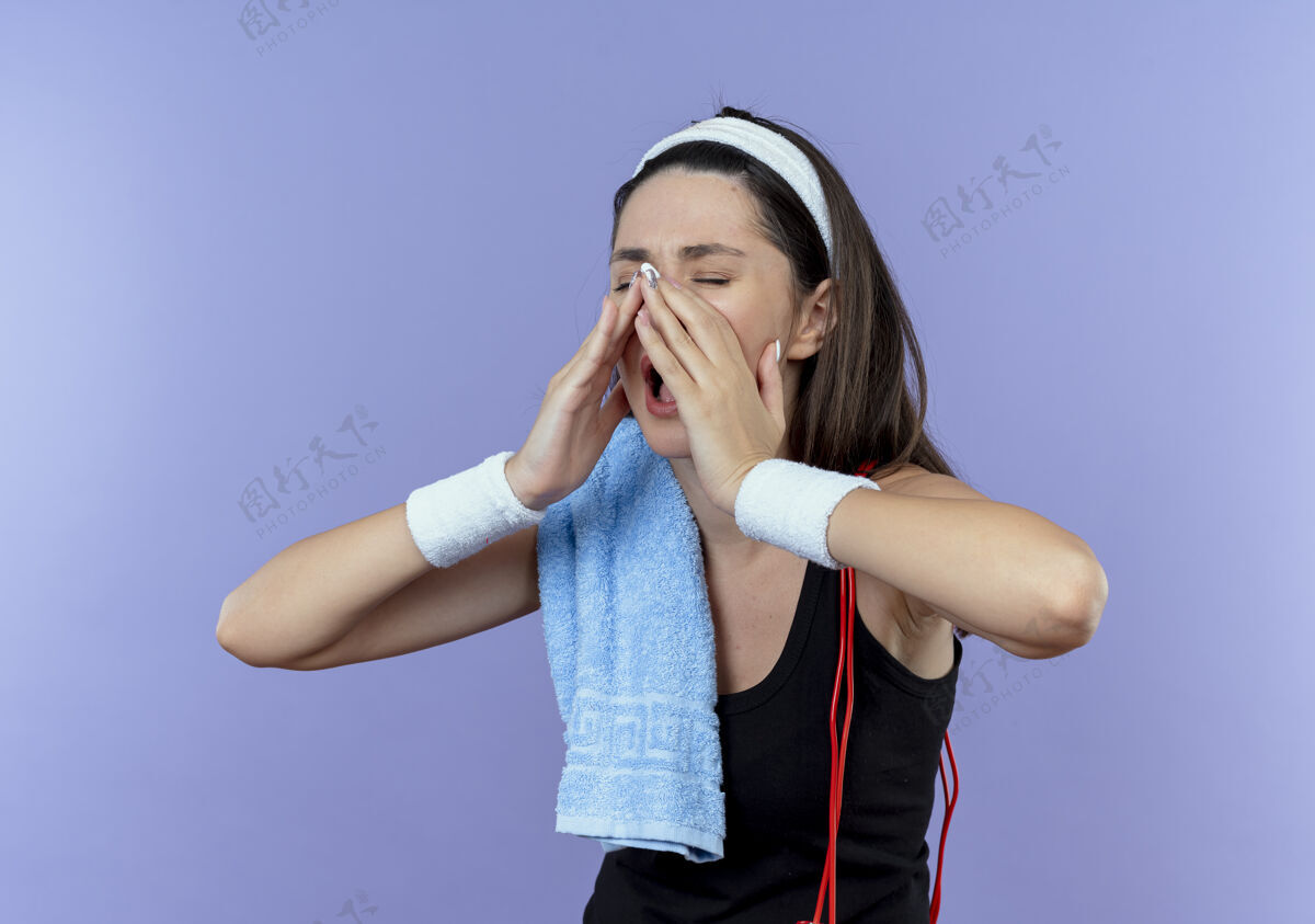 健身戴着头巾 肩上扛着毛巾的年轻健身女士站在蓝色背景下 用手靠近嘴巴喊着或叫着别人肩膀头带毛巾
