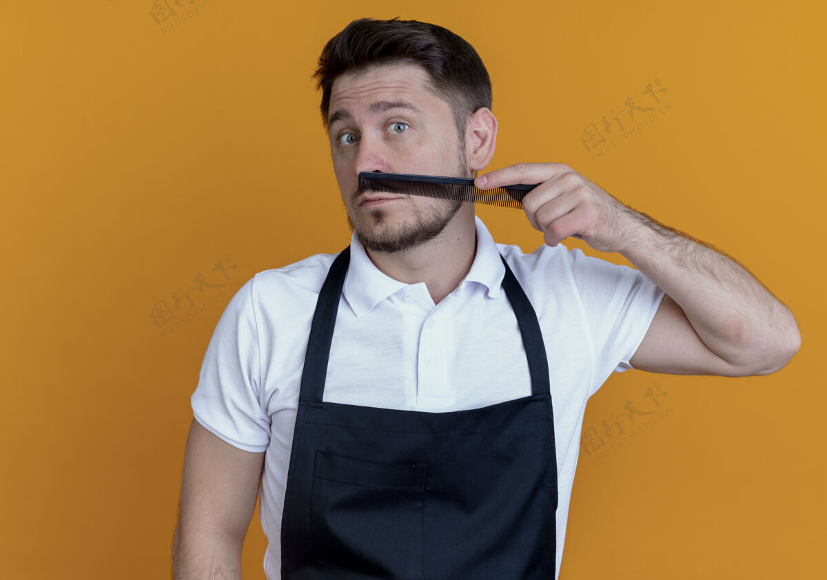 围裙围裙上的理发师梳着胡子 站在橙色背景上看着摄像机梳子胡子理发师