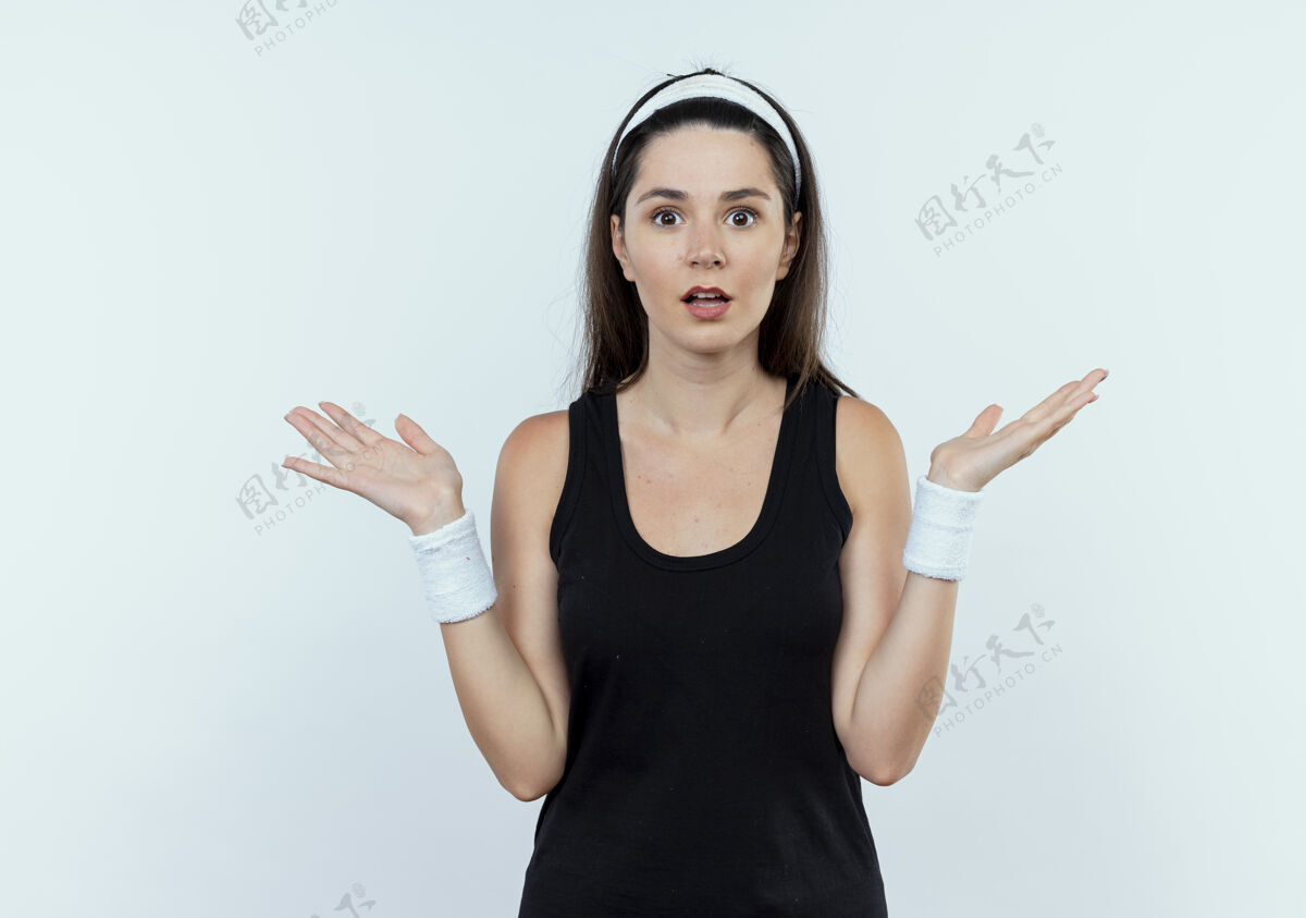 相机戴着头巾的年轻健身女看着镜头迷茫而不确定地伸开双臂站在白色背景上壁板手臂头带