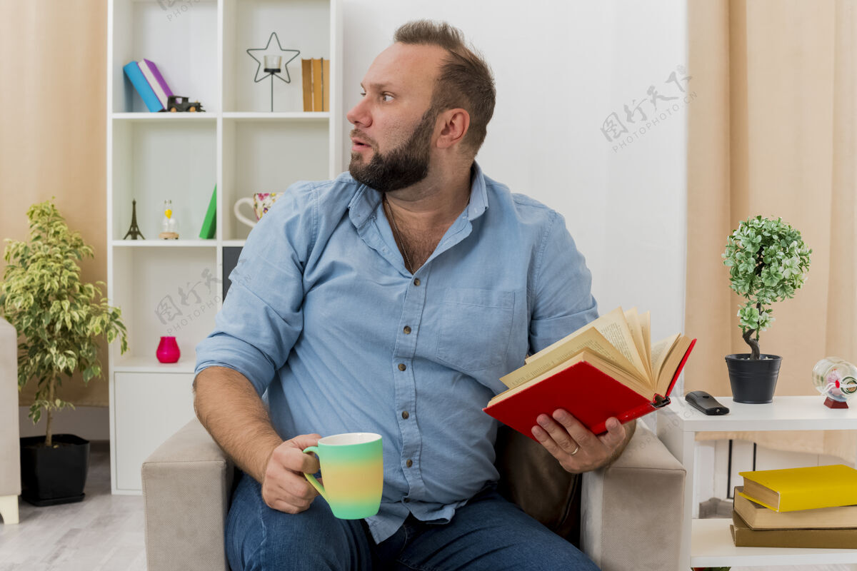 扶手椅焦虑的成年斯拉夫人坐在扶手椅上 手里拿着书和杯子 看着客厅里的一边男人壁板客厅