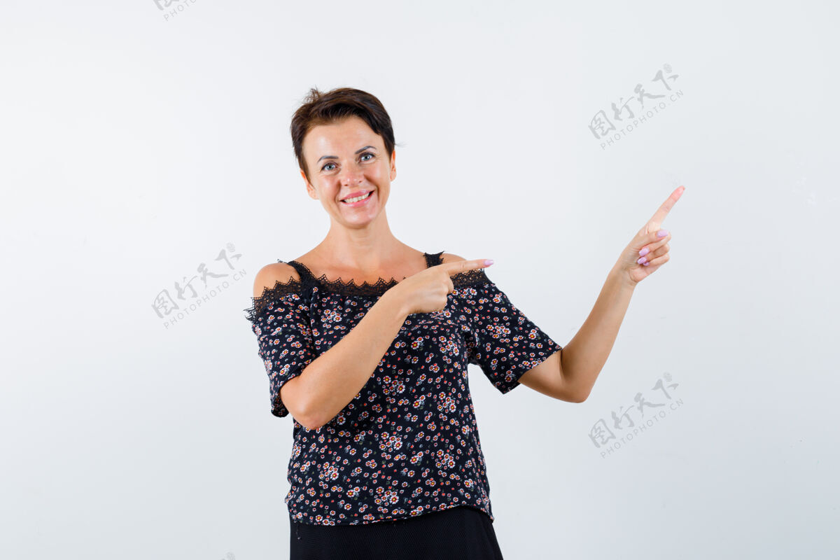 商务成熟的女人用食指指着右边 穿着花衬衫 黑色裙子 看起来很快乐 正面视图成熟姿势目录