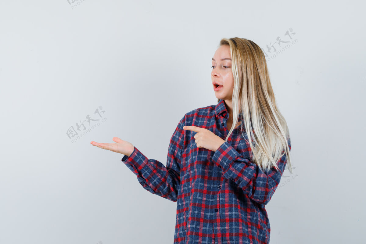 焦点金发女人举起手拿着什么东西 用另一只手指着它 穿着格子衬衫 看起来很专注 前视图提高健康拿着