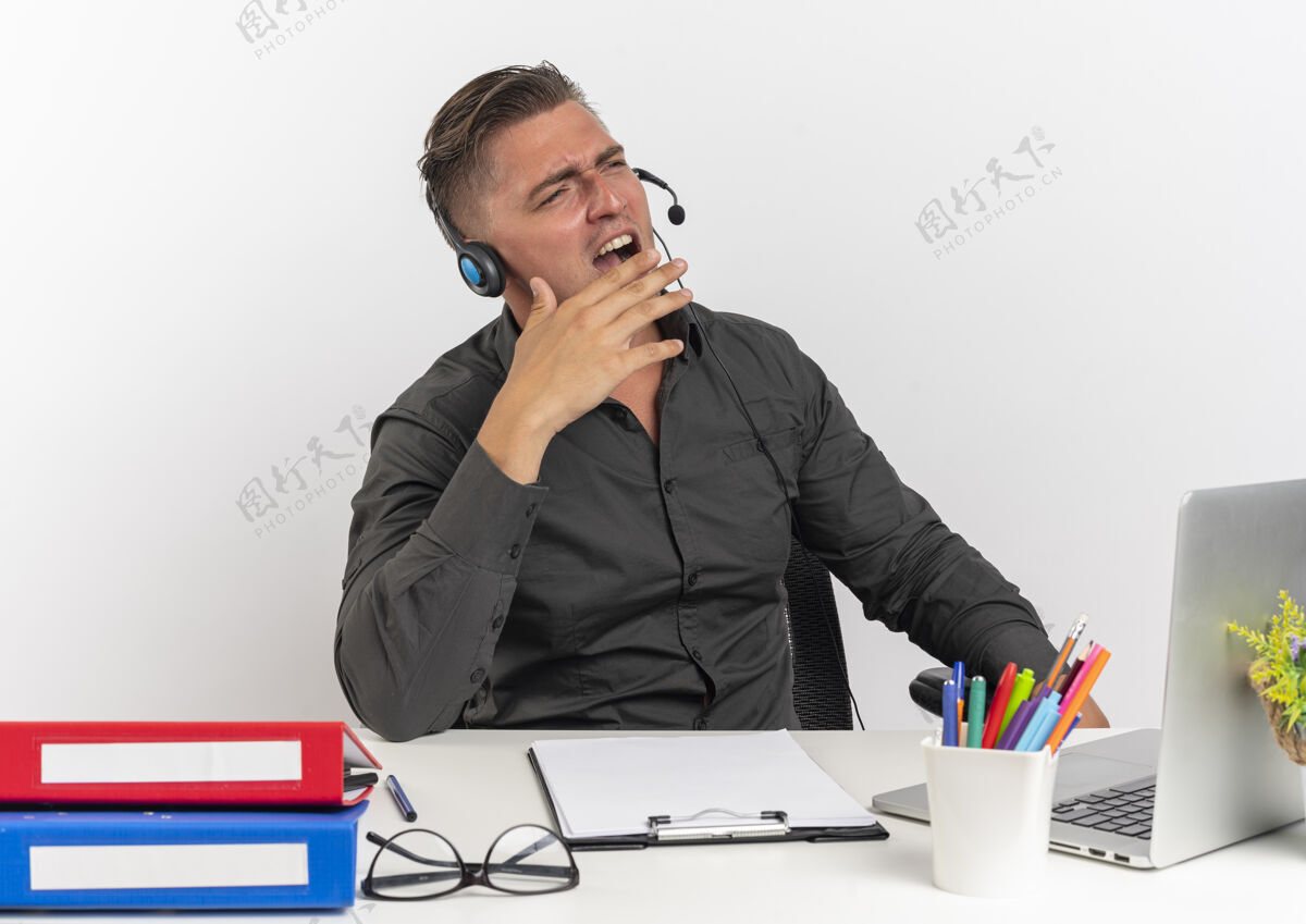 年轻人年轻恼怒的金发上班族戴着耳机坐在办公桌旁 拿着办公工具 用笔记本电脑对着一个人大喊大叫 他看着一个被隔离在白色背景上的有复印空间的人工作人员耳机侧身