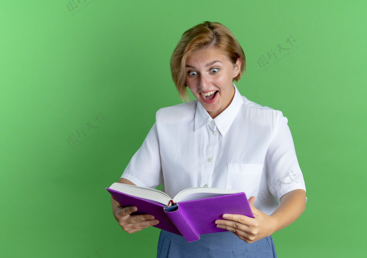 女孩年轻惊讶的金发俄罗斯女孩看着书在绿色背景与复制空间隔离金发女郎年轻俄国人