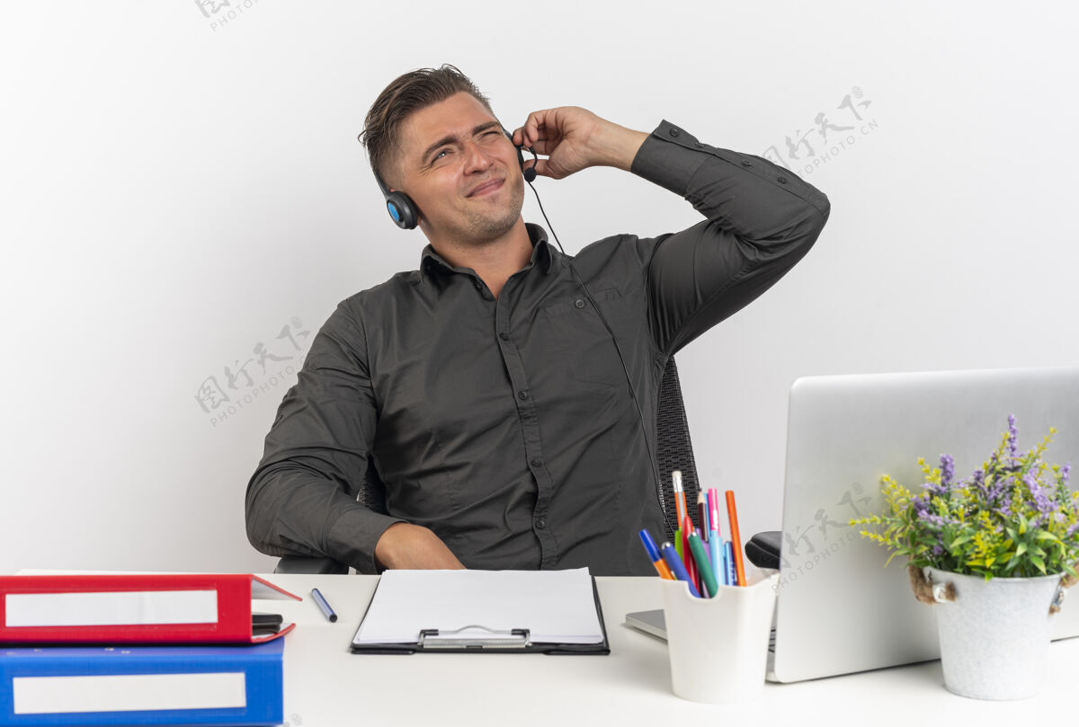 拿着年轻恼怒的金发上班族戴着耳机坐在办公桌旁 拿着办公工具 用笔记本电脑拿着耳机 试图在白色背景和复印空间中听到孤立的声音坐着使用耳机