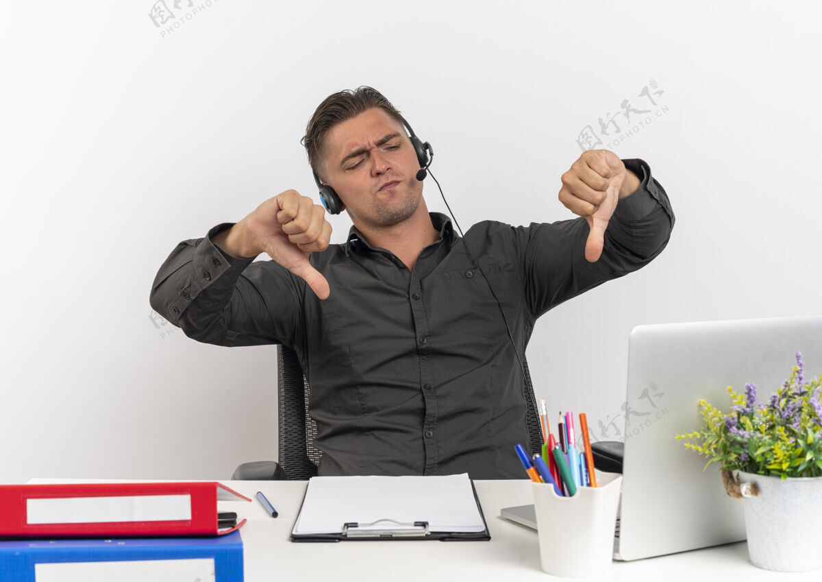 拇指年轻恼怒的金发上班族戴着耳机坐在办公桌旁 拿着办公工具 看着笔记本电脑 大拇指朝下 隔离在白色背景上 留着复印空间工作人员年轻金发