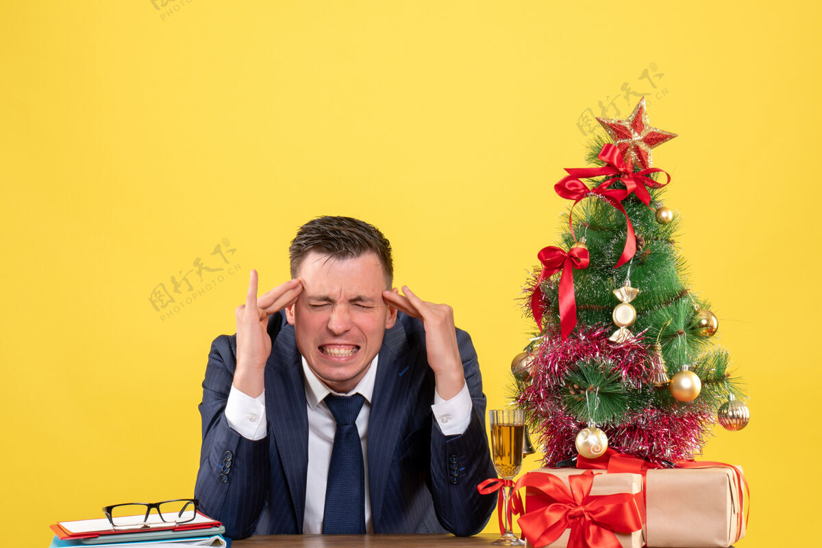 礼物前视图愤怒的男人把手指放在他的太阳穴上 坐在圣诞树旁的桌子上 黄色背景上的礼物坐西装太阳穴