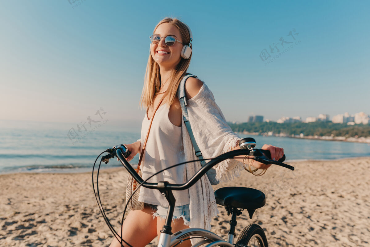 放松年轻迷人的金发微笑的女人 戴着耳机骑着自行车走在沙滩上听音乐女性阳光旅行者