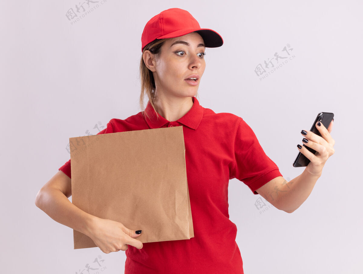 纸穿着制服的年轻漂亮的女送货员拿着纸包 看着隔离在白墙上的电话制服举行人