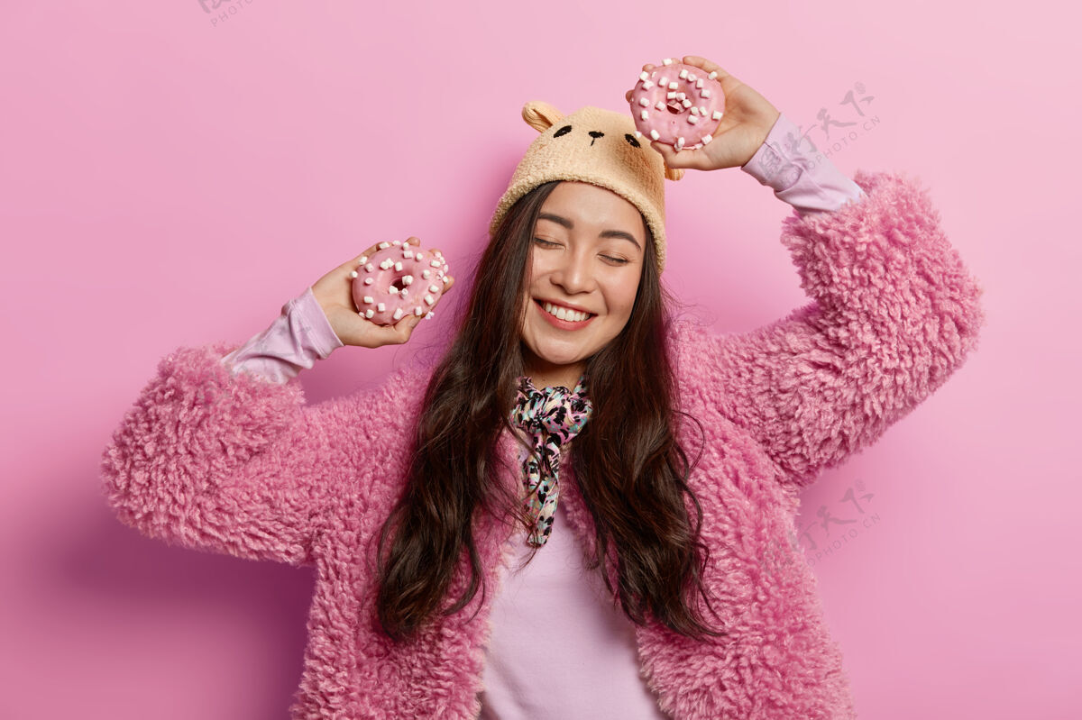甜甜圈无忧无虑的女士拿着两个甜甜圈快乐地跳舞 在室内玩得很开心 穿着粉色外套 戴着棕色帽子蛋糕釉面美味