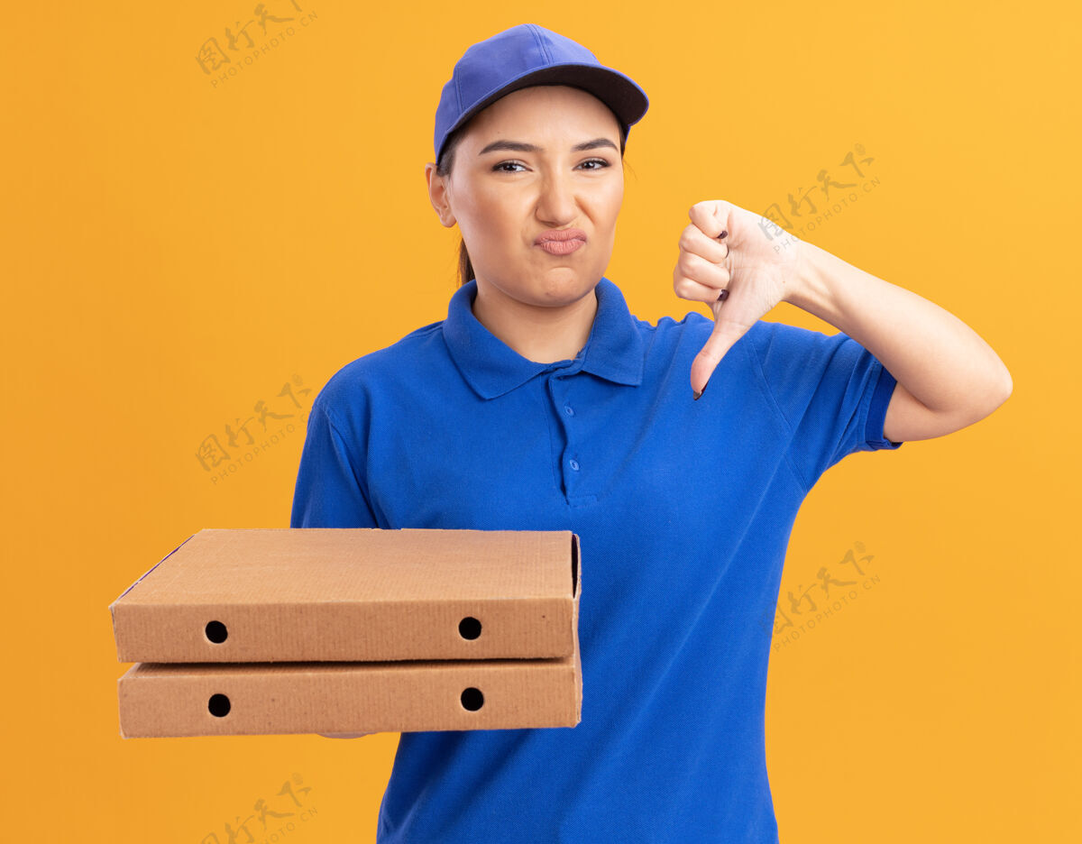 站着不满意的年轻女送货员穿着蓝色制服 戴着帽子 拿着披萨盒 不高兴地站在橙色的墙上竖起大拇指看着前面放下制服年轻