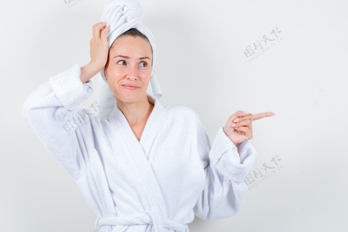 女性身穿白色浴袍的年轻女士 手扶毛巾头 同时指向右侧 神情愉悦 俯视前方新鲜护理人