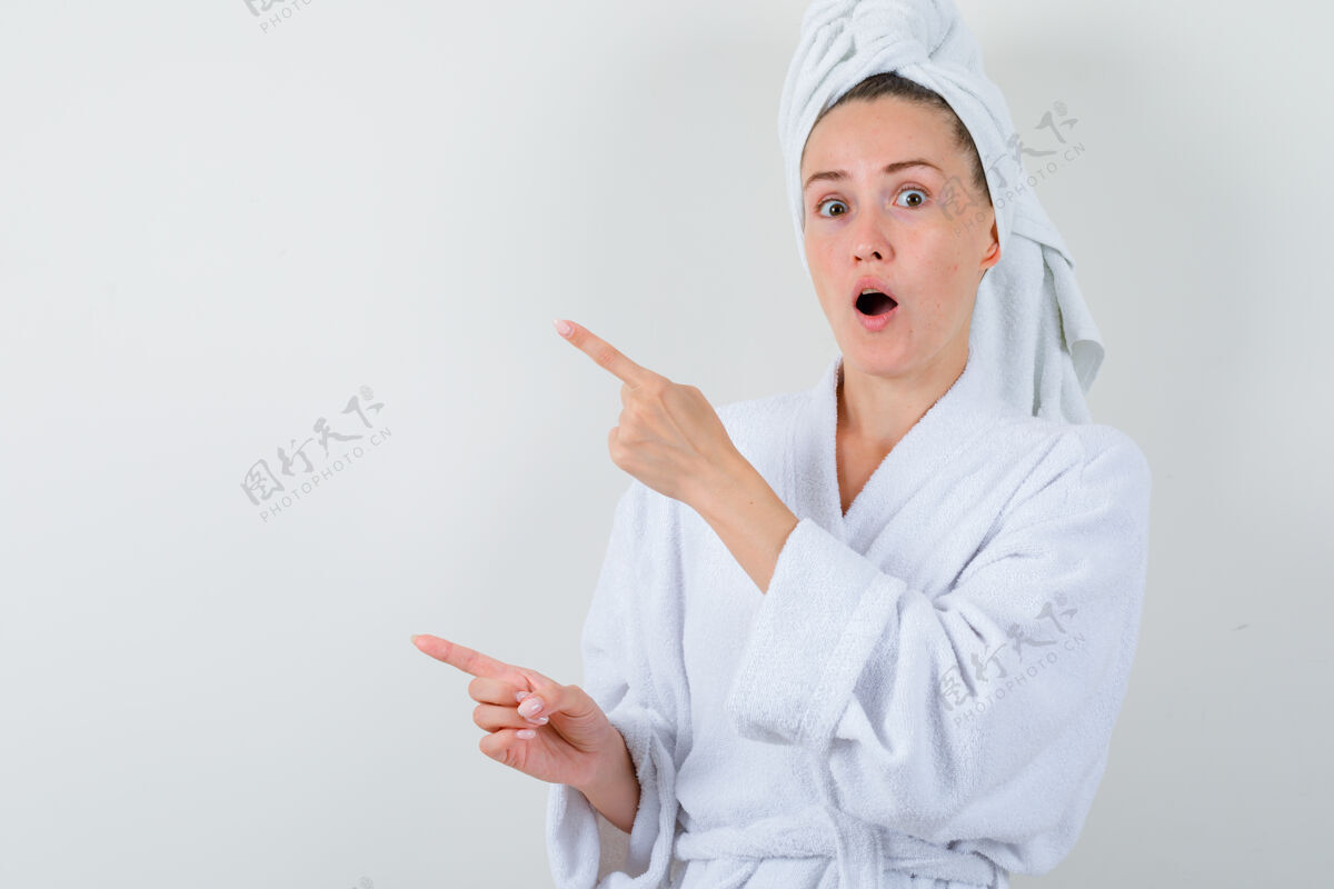 微笑年轻女士指着旁边的白色浴衣 毛巾和期待惊讶 前视图完美黑发年轻