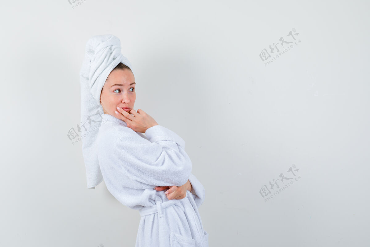 漂亮一个年轻的女人 手放在下巴上 穿着白色浴衣 拿着毛巾 神情好奇地看着别处乐趣女人好奇