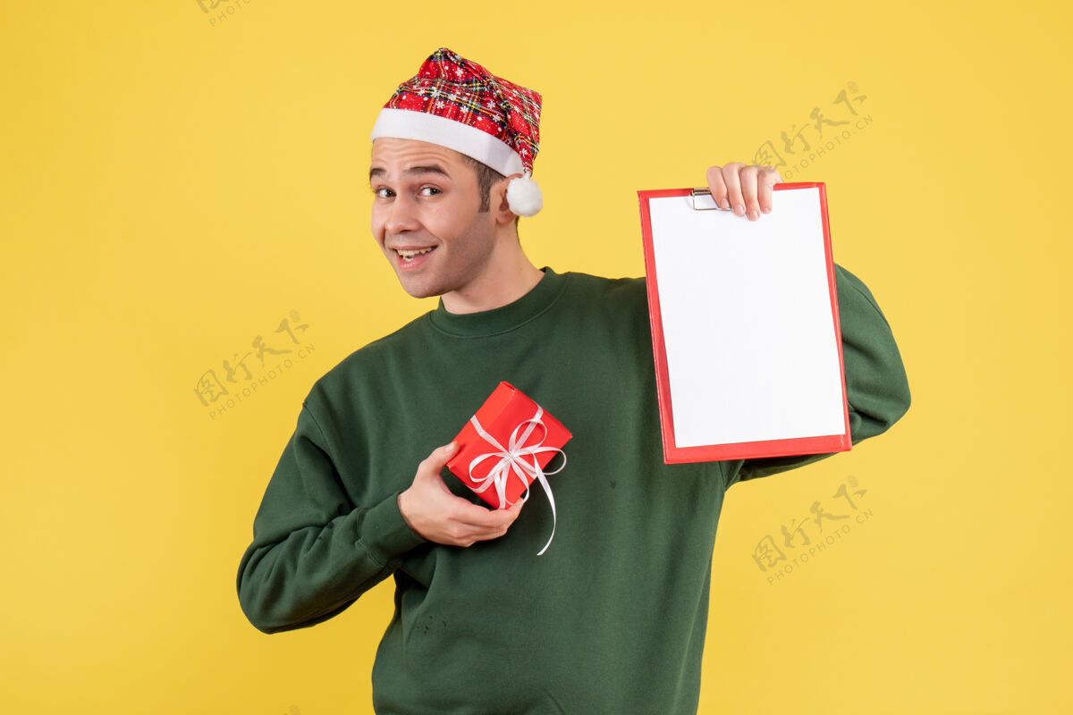 圣诞节正面图：戴着圣诞帽的年轻人拿着剪贴板和礼物站在黄色背景上持有建筑背景