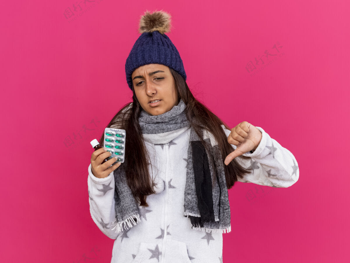 帽子未出狱的年轻病女孩戴着冬天的帽子 戴着围巾 拿着玻璃瓶里的药 拇指朝下 隔离在粉色背景上向下不愉快药物