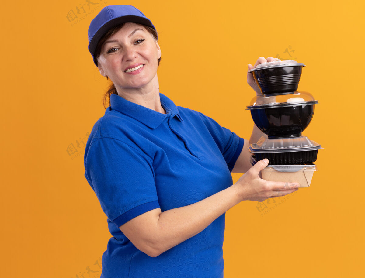 女人身穿蓝色制服 头戴鸭舌帽的中年女送货员站在橙色的墙上 手里拿着一叠食品包 面带微笑地看着前面叠着欢呼盒子