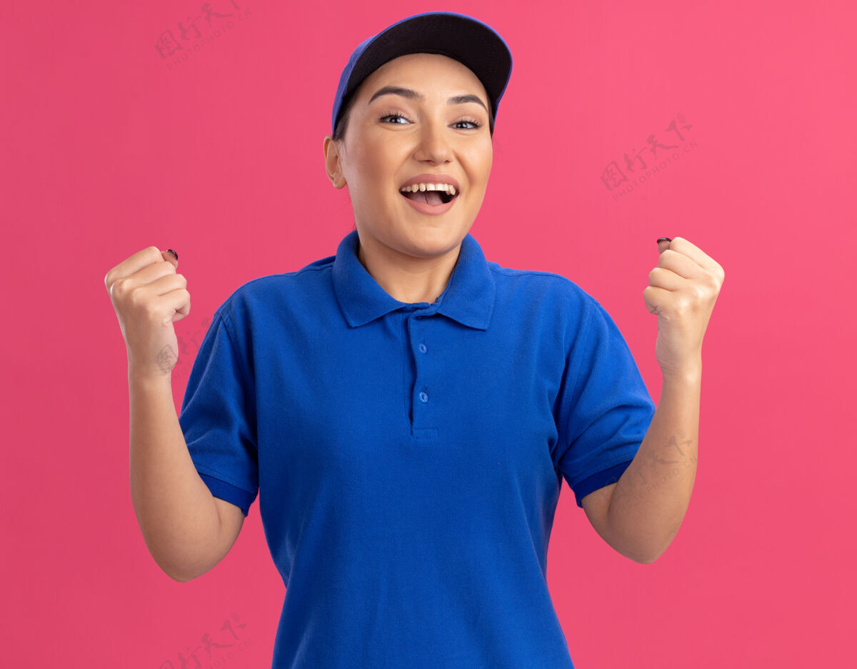快乐身穿蓝色制服 头戴鸭舌帽的年轻女送货员站在粉红色的墙上 高兴而兴奋地看着前面紧握的拳头帽子拳头兴奋