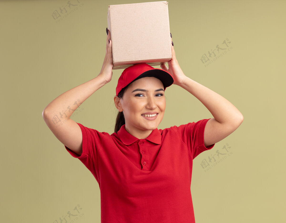 纸板身穿红色制服 头戴鸭舌帽的年轻女送货员举着纸板箱 站在绿色的墙上 面带微笑地看着前面女人年轻人欢呼
