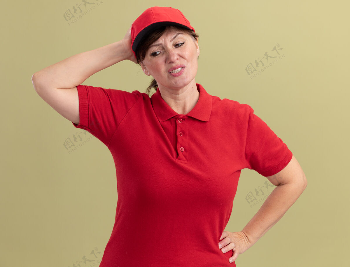 手身穿红色制服 戴着帽子的中年女送货员 站在绿色的墙壁上 看起来很困惑 因为她站错了头错误头立场