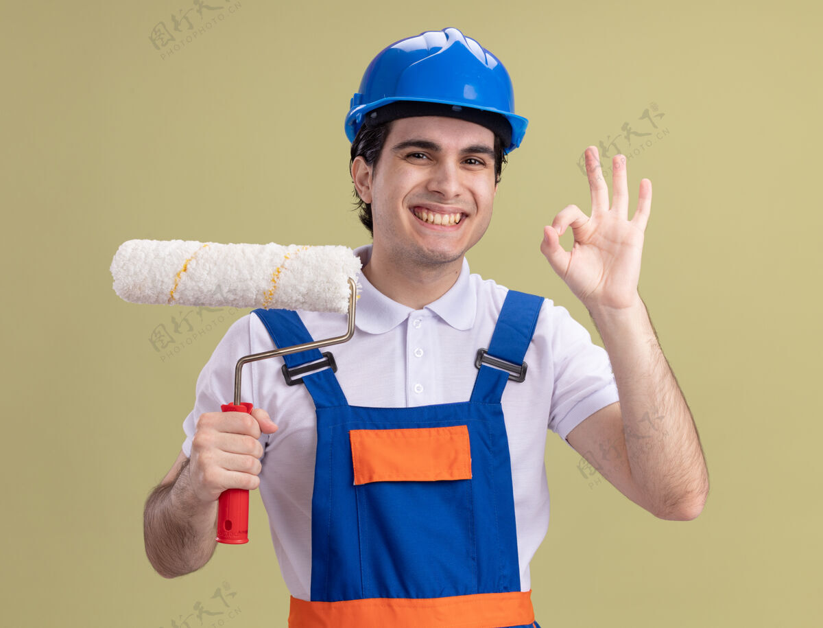 建筑工人年轻的建筑工人穿着建筑制服 戴着安全帽 拿着油漆辊 面带微笑地看着前面 绿色的墙上挂着“ok”的牌子微笑滚轴年轻
