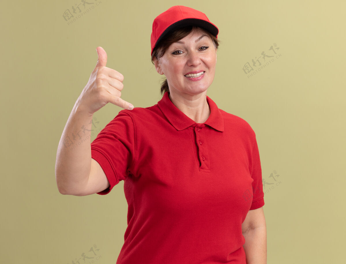 欢呼身穿红色制服 头戴帽子的中年送货妇女站在绿色的墙上 面带微笑地做着叫我的手势送货手势站立