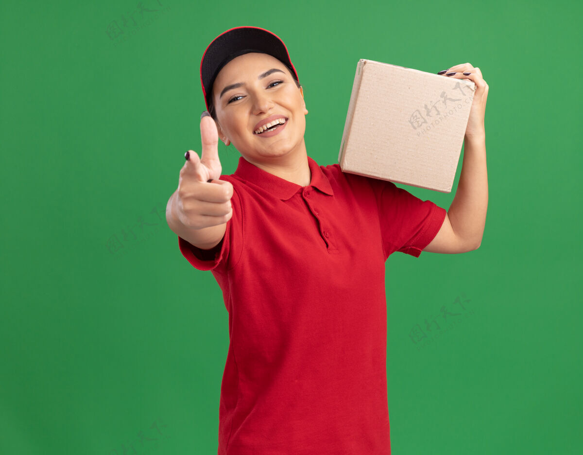 目录身着红色制服 戴着帽子 拿着纸板箱 食指指着前面 快乐地站在绿色墙壁上的年轻送货员女人递送年轻
