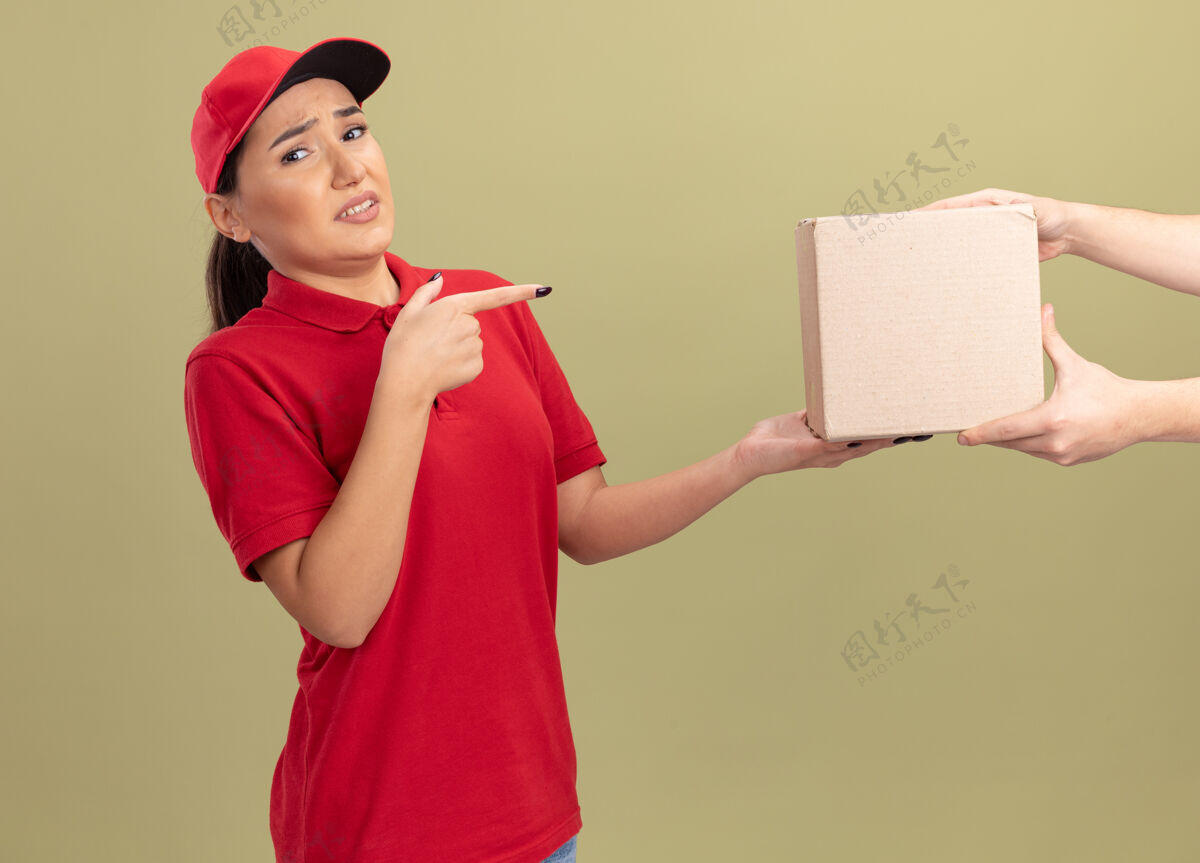 目录身着红色制服 戴着帽子的年轻送货员在收包裹时感到困惑 食指指着站在绿色墙上的盒子制服交货包装