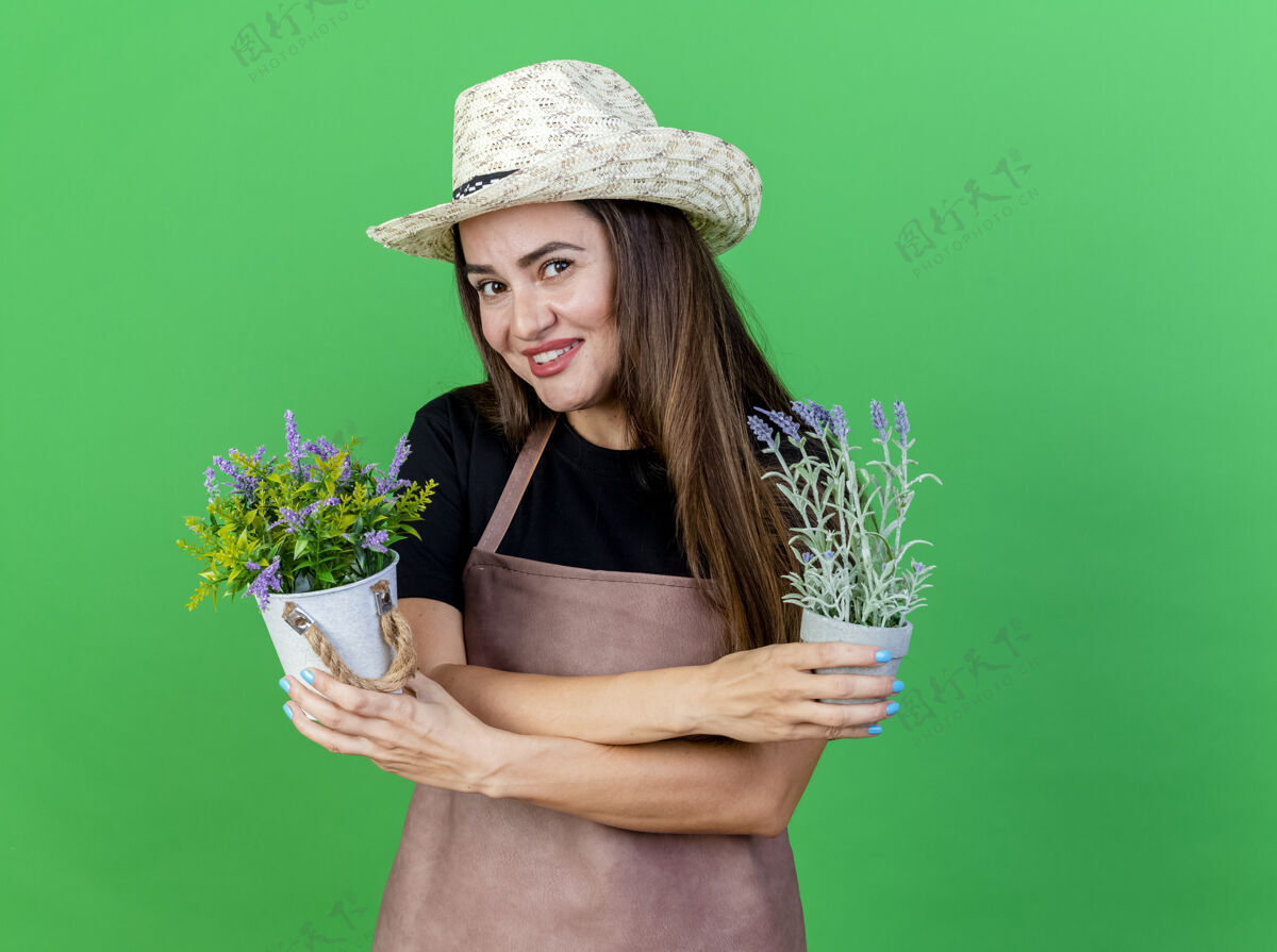 园艺微笑美丽的园丁女孩穿着制服 戴着园艺帽 在绿色的背景下 在花盆里拿着和交叉的花花盆花女孩