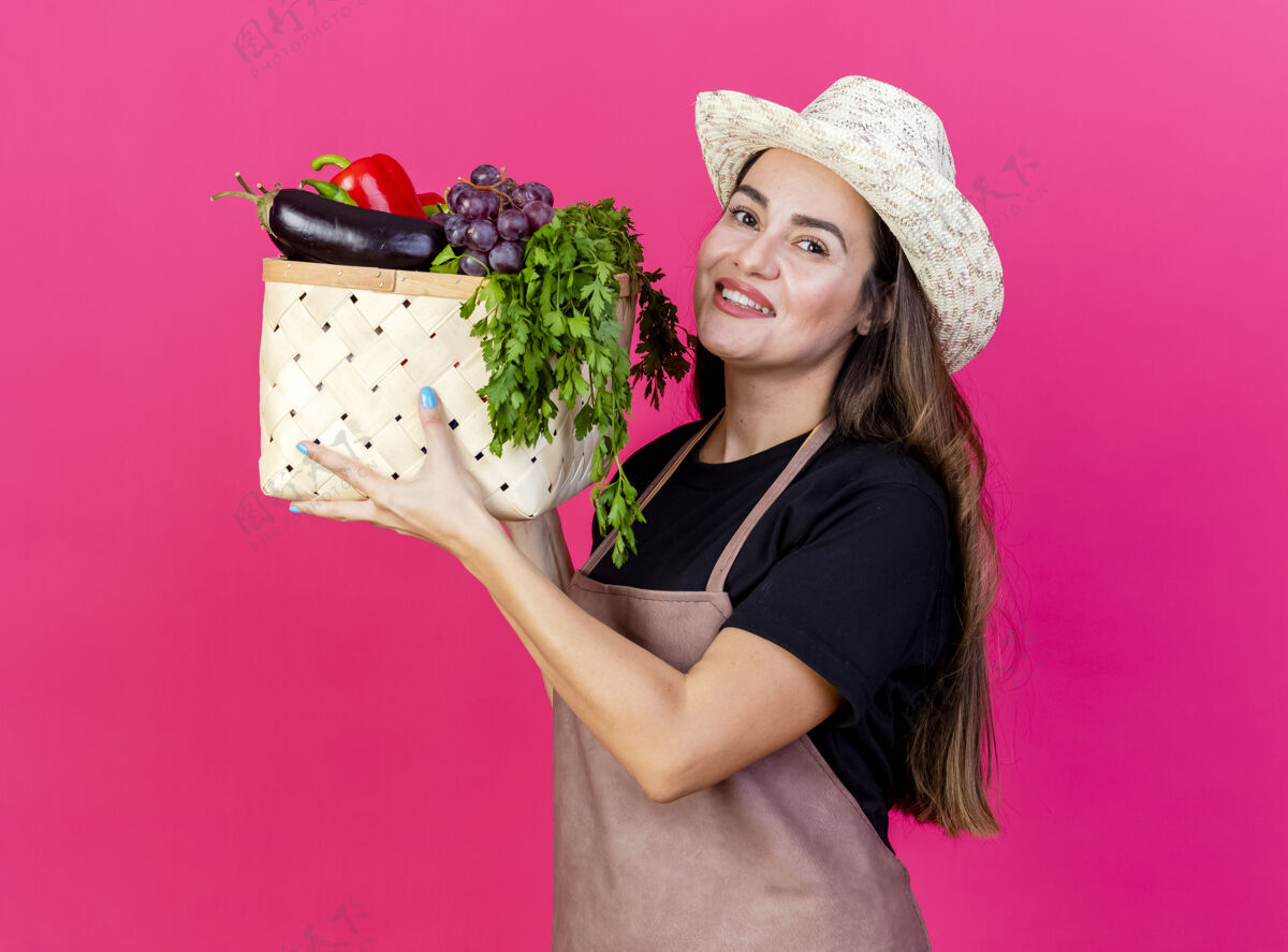 帽子微笑美丽的园丁女孩穿着制服 戴着园艺帽 在粉红色的背景上孤立地举起菜篮子制服穿篮子