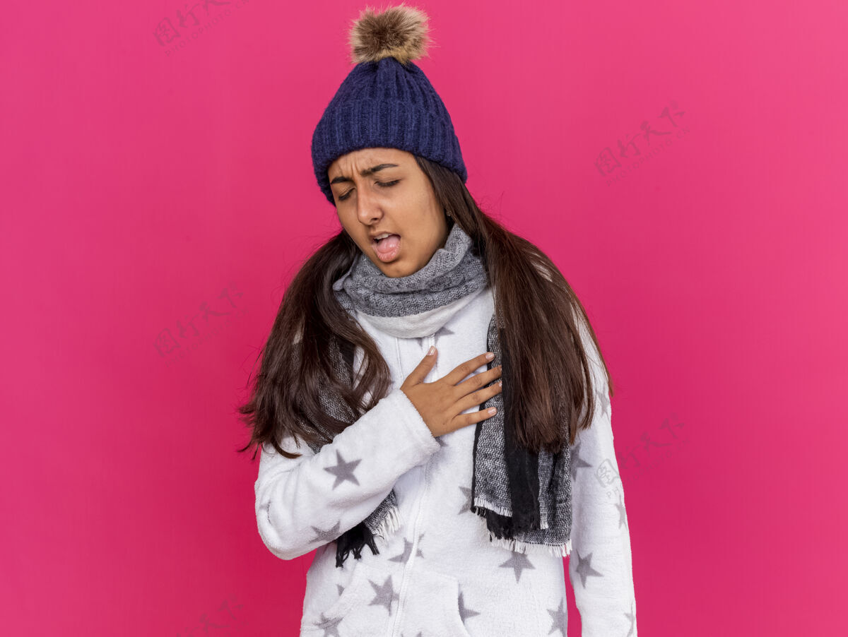 女孩咳嗽的年轻病女孩闭着眼睛戴着冬天的帽子戴着围巾把手放在心上隔离在粉红色的背景上咳嗽疾病围巾