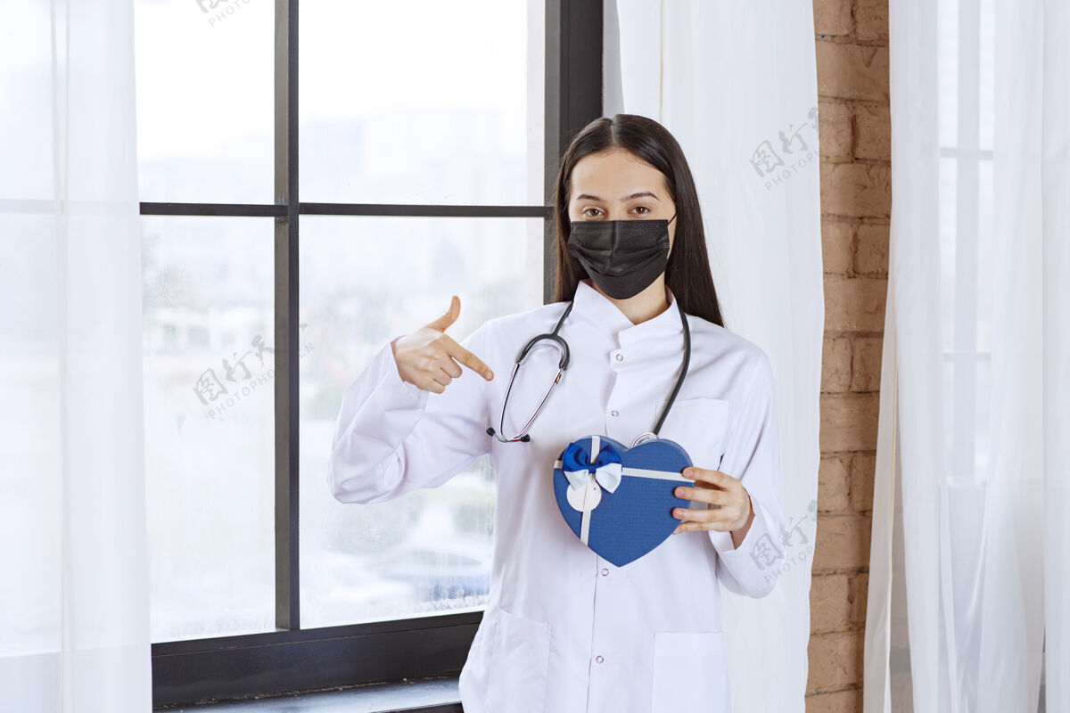 女性医生拿着听诊器和黑色口罩 手里拿着一个蓝色心形礼盒病毒医院治疗