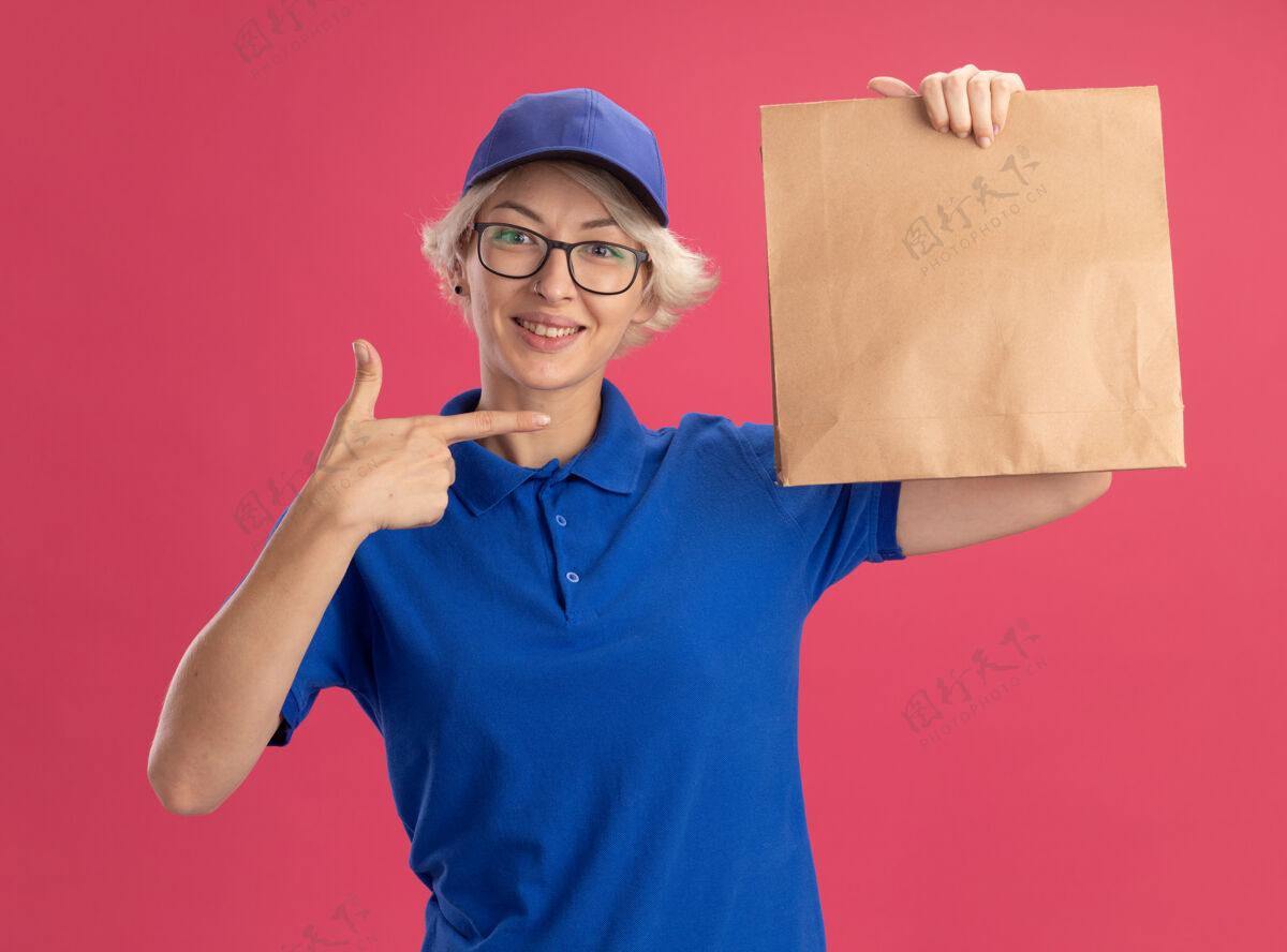帽子身穿蓝色制服 戴着眼镜的年轻女送货员手里拿着纸包裹 兴高采烈地笑着 用食指指着粉色墙上的包裹手指戴着指点符号